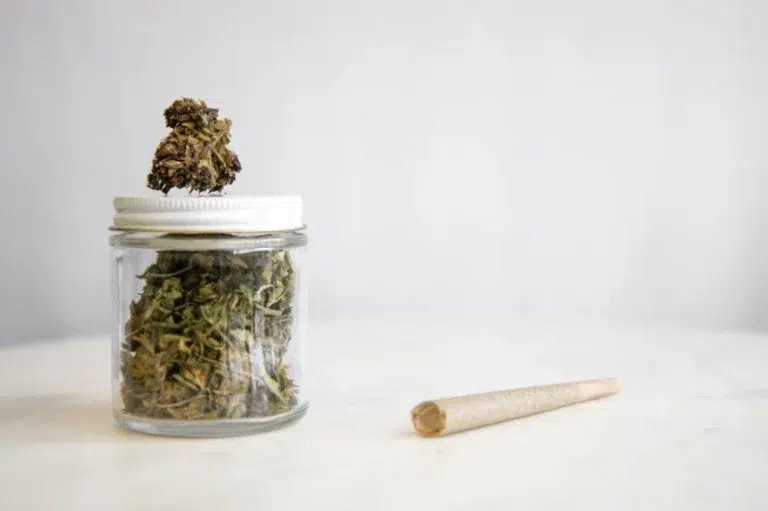 A jar filled with cannabis with a bud in top, plus a weed joint laying on the table.